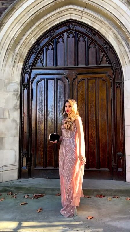 Roopsi Bansal in a rose gold beaded column gown with a cape, standing in front of a grand arched wooden door