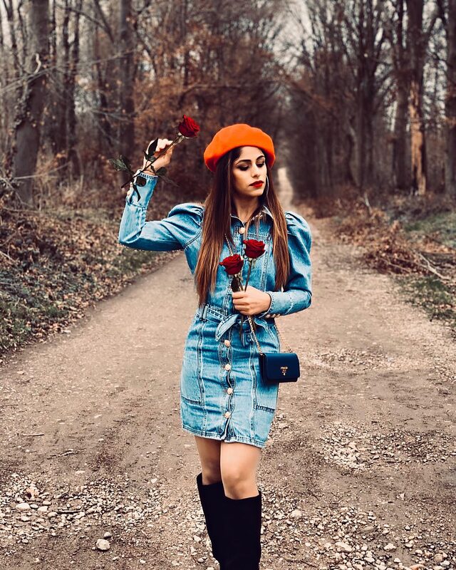 Standing on a dirt path through bare winter trees, wearing a denim dress with puff sleeves and an orange beret, holding two red roses, with a blue Prada bag