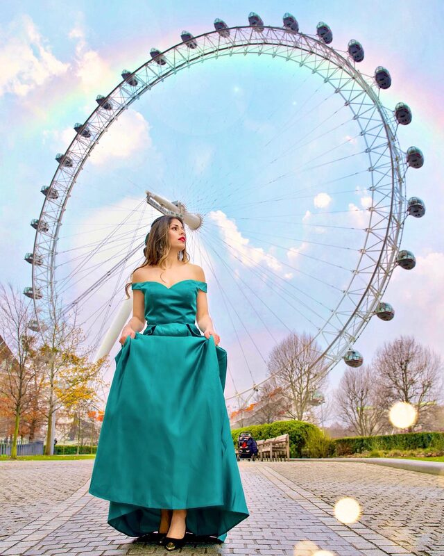 Roopsi Bansal in a teal off-shoulder satin ball gown with a full skirt, standing in front of a large Ferris wheel
