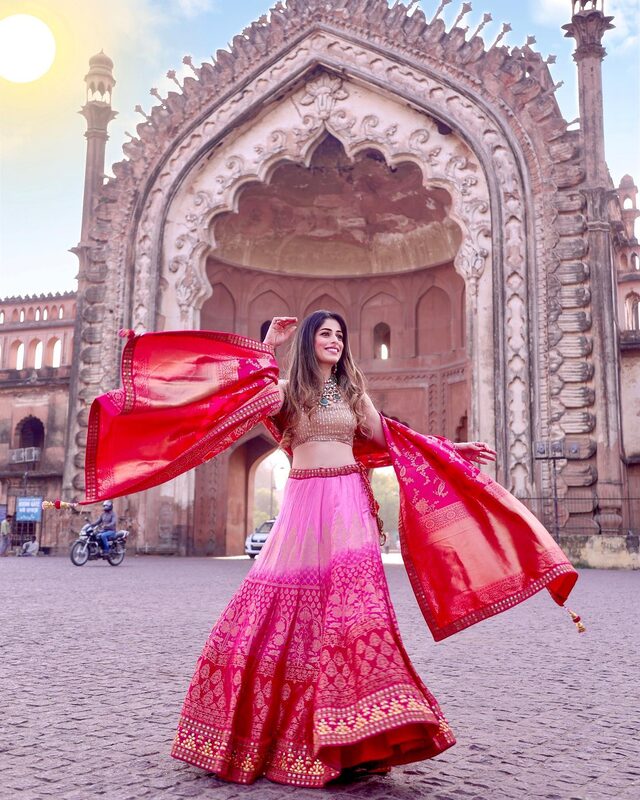Roopsi Bansal twirling in an ombre pink and magenta lehenga with a red banarasi dupatta in front of the towering Rumi Darwaza gateway in Lucknow