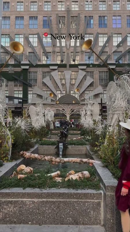 Roopsi Bansal in a burgundy poncho and wide-brim hat facing Saks Fifth Avenue's art deco holiday facade in New York