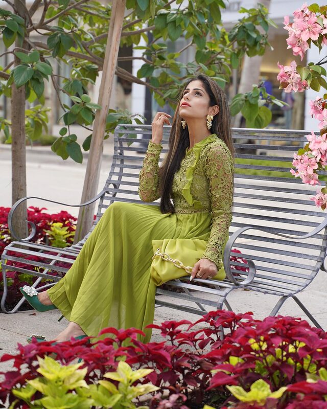 Roopsi Bansal in a green lace and chiffon midi dress seated on a garden bench surrounded by red poinsettias and pink blossoms