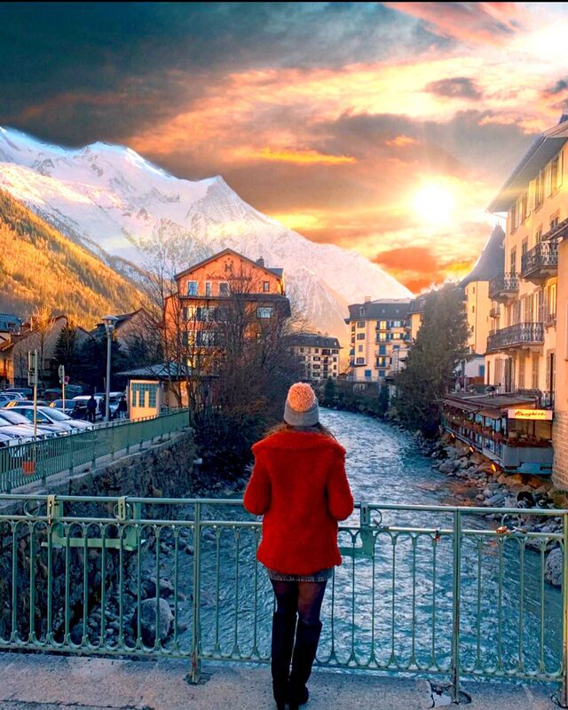 Standing on the same green iron bridge in Chamonix as before, now in a fuzzy red coat and cream pom-pom beanie, with a dramatic orange-pink sunset lighting up the sky over the river and mountains