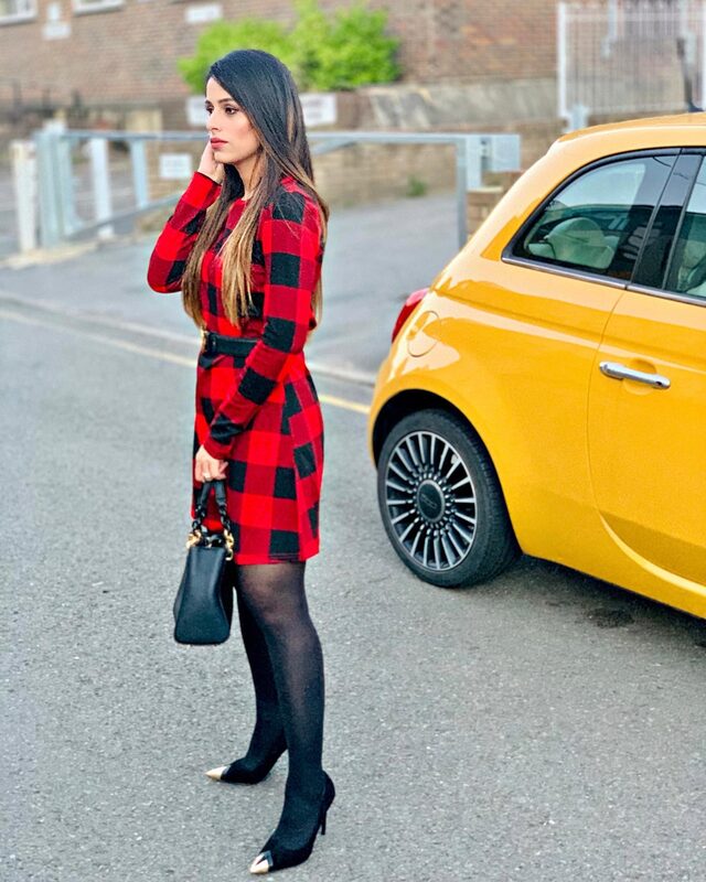 Roopsi Bansal in a red and black plaid dress and pointed heels, standing on a London street beside a bright yellow Fiat