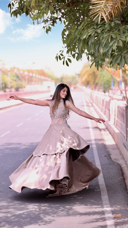 Roopsi Bansal twirling with arms outstretched in a pastel taupe embellished gown, on an empty tree-lined road
