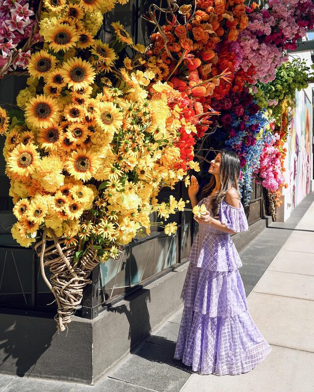 Roopsi Bansal in a lilac off-shoulder windowpane tiered ruffle dress from SHEIN, reaching toward a wall covered in sunflowers, orange roses, and pink blooms in Buckhead Village