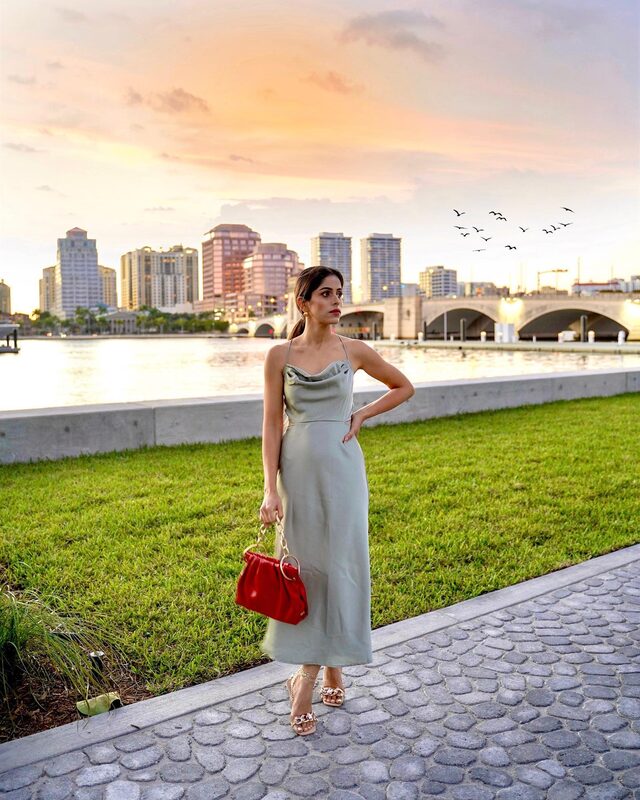 Roopsi Bansal in a sage green cowl-neck slip dress, holding a red handbag, standing on a waterfront path with the West Palm Beach skyline glowing at sunset behind her
