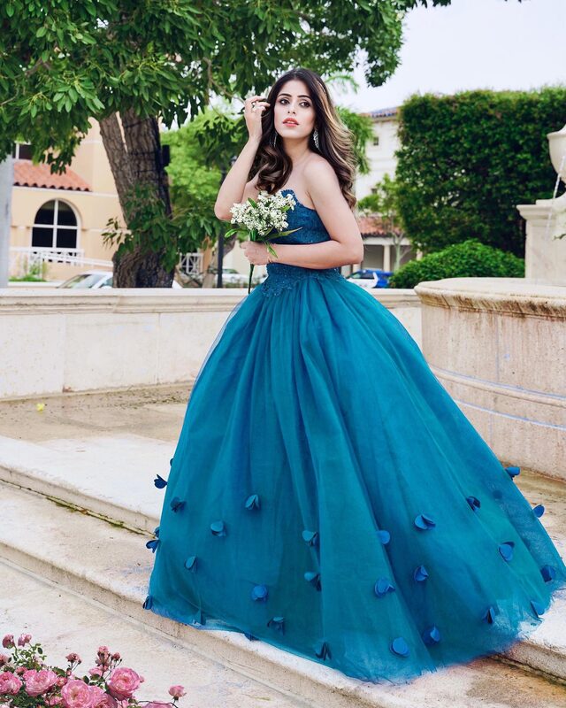 Roopsi Bansal in a teal strapless ball gown covered in petal appliqués, holding a small bouquet in front of a stone fountain