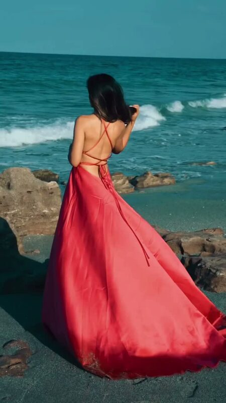 Roopsi Bansal from behind in a coral red backless gown from JJ's House, walking along a rocky Florida beach toward the ocean