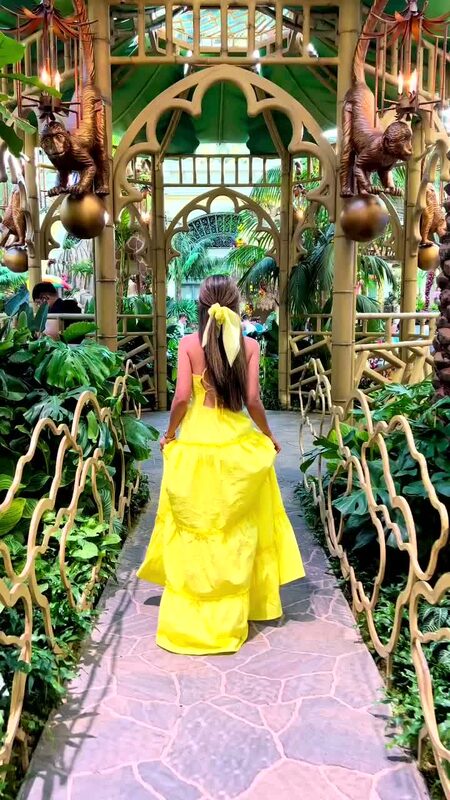 Roopsi Bansal from behind in a yellow tiered dress, walking through a golden gazebo with monkey sculptures at the Bellagio Conservatory in Las Vegas