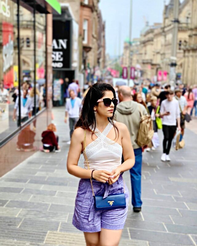 Walking a busy Scottish shopping street wearing a white crochet halter top, lavender linen shorts, sunglasses, and a navy Prada crossbody bag