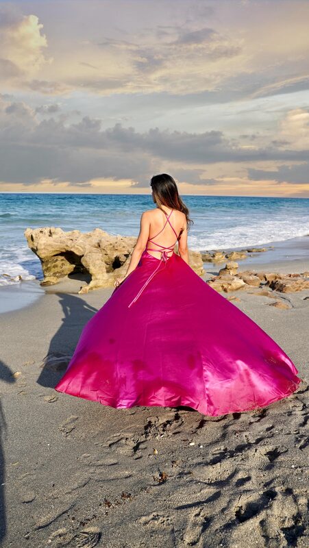 Roopsi Bansal in a magenta satin A-line prom gown with a sweep train from JJ's House, back to the camera, standing on the rocks at Coral Cove Park with waves rolling in