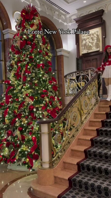 Roopsi Bansal in a red and white cropped sweater set on the grand staircase of Lotte New York Palace, decorated with a large Christmas tree