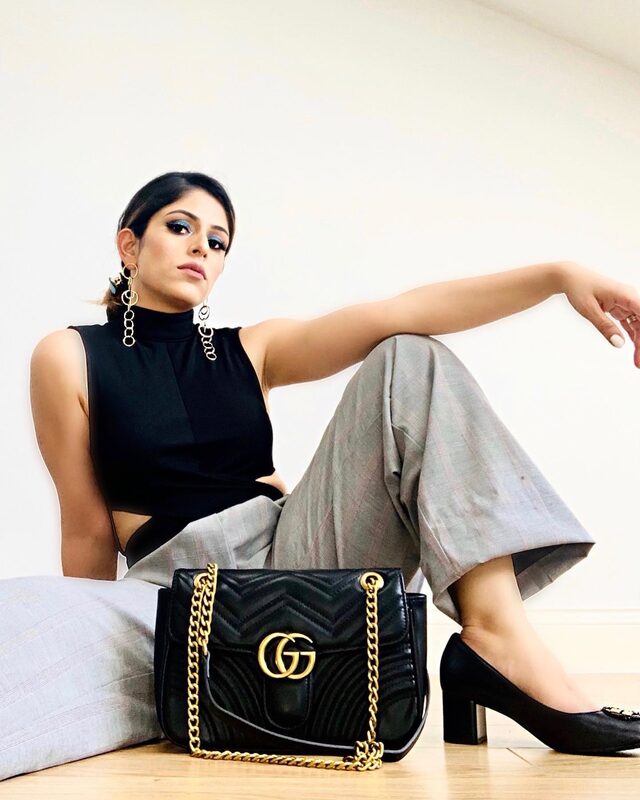 Sitting on the floor against a couch wearing a black cutout top and grey plaid wide-leg trousers, gold drop earrings, with a black Gucci Marmont bag on the floor in front