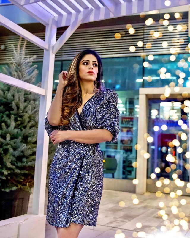 Roopsi Bansal in a silver-blue sequin wrap blazer dress at night, standing under a white pergola with a curtain of golden bokeh lights behind her