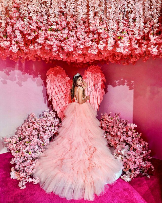 Roopsi Bansal in a blush tulle ball gown, tiara, and large pink feathered angel wings, standing beneath a canopy of hanging cherry blossoms at a floral installation
