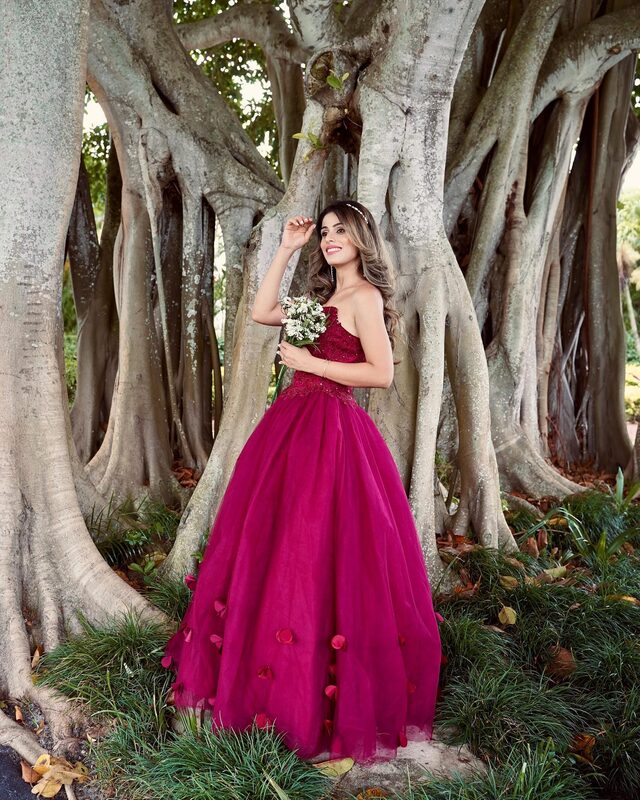 Roopsi Bansal in a magenta strapless ball gown covered in petal appliqués, holding a small bouquet of wildflowers among the roots of old banyan trees