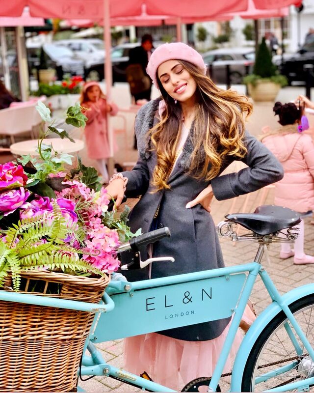 Roopsi Bansal in a pink beret and grey fur-collared coat, posing beside a teal bicycle with a flower basket outside El&N London cafe