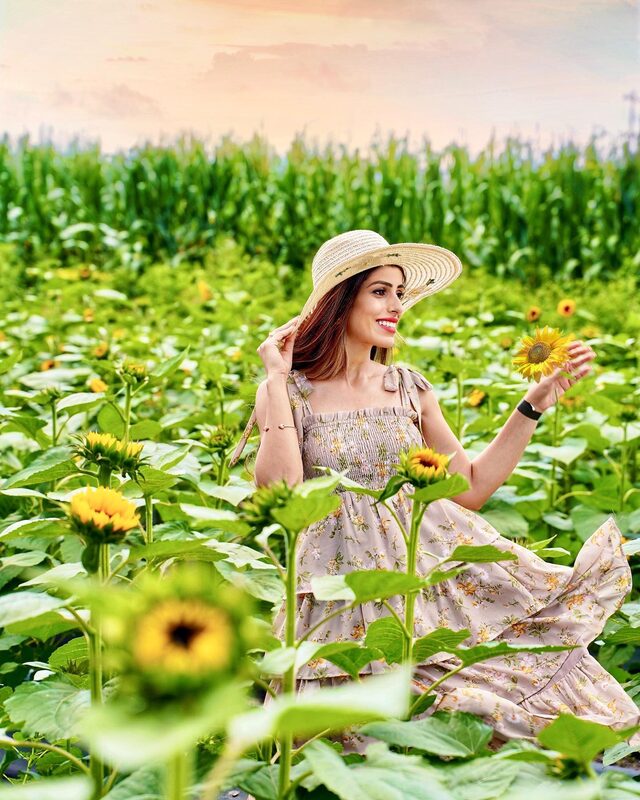 Roopsi Bansal in a straw hat and tiered floral dress, holding a sunflower in a sunflower field at Bedner's Farm, Florida