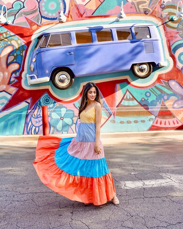 Roopsi Bansal in a yellow, blue, pink, and orange colorblock ruffle-hem cami dress from SHEIN, standing in front of a psychedelic mural of a VW bus at Mellow Mushroom, Atlanta