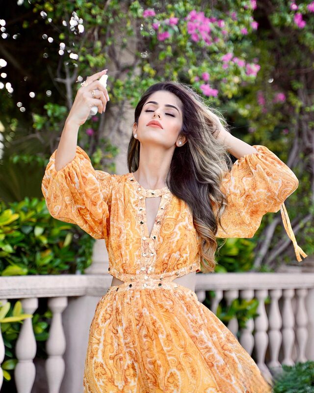 Roopsi Bansal in an orange paisley-print dress with waist cutouts, spritzing facial spray with her eyes closed in front of a wall of pink bougainvillea