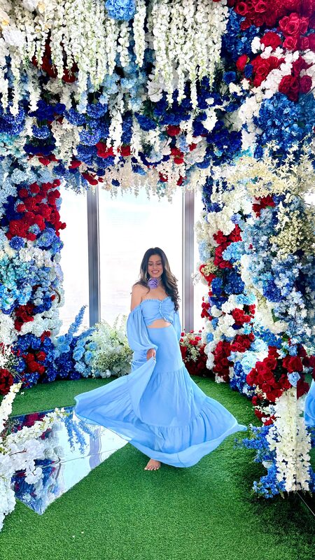 Roopsi Bansal in a powder blue off-shoulder dress twirling inside a red, white and blue floral archway installation at The Edge, Hudson Yards