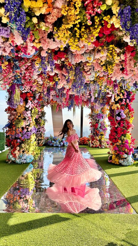 Roopsi Bansal in a pink polka-dot tiered dress standing under a rainbow flower archway with a mirrored floor at The Edge's Sky Bloom installation