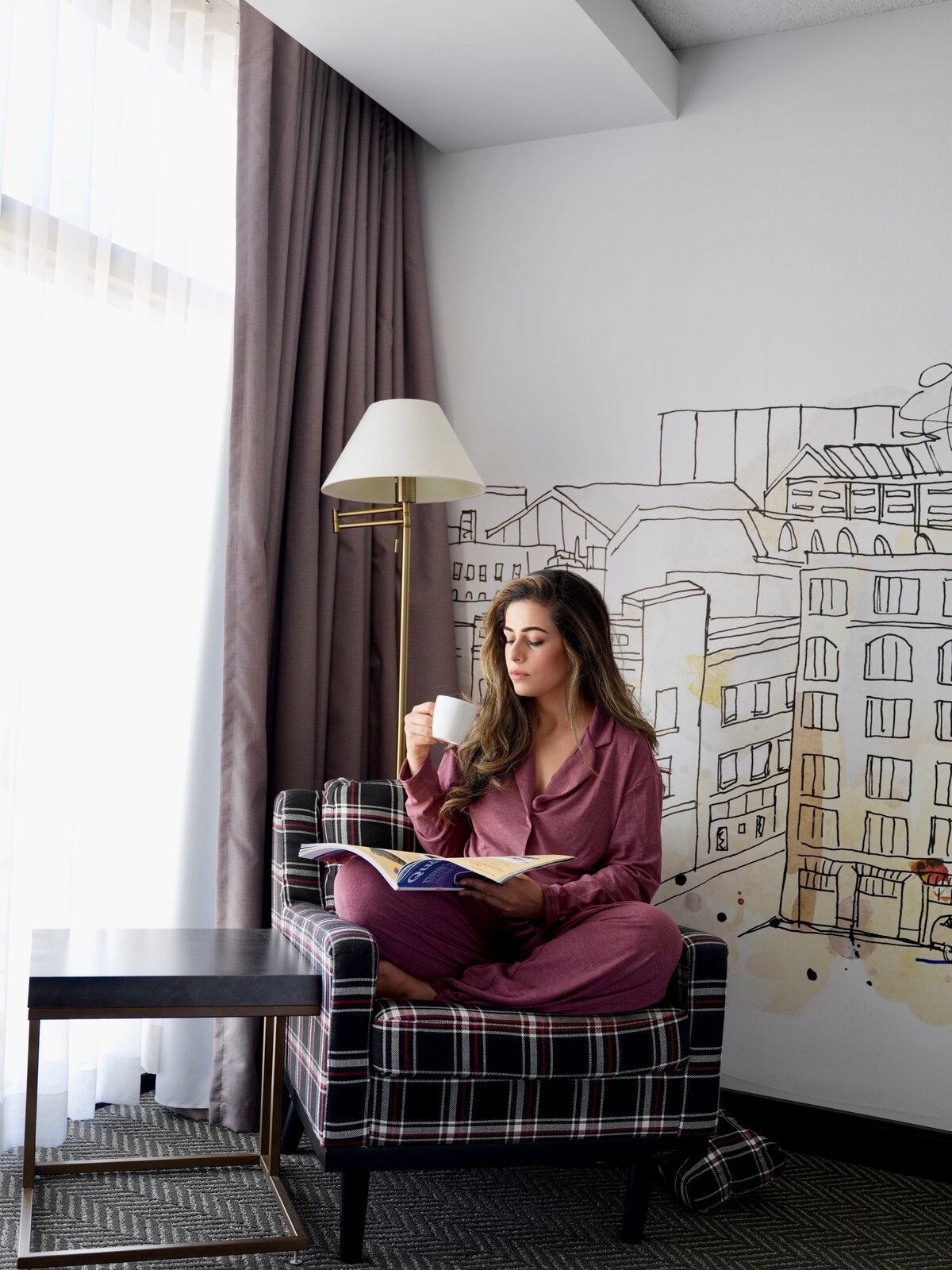 Roopsi Bansal in a mauve pajama set sitting in a plaid armchair by a hotel window, drinking coffee and reading a magazine