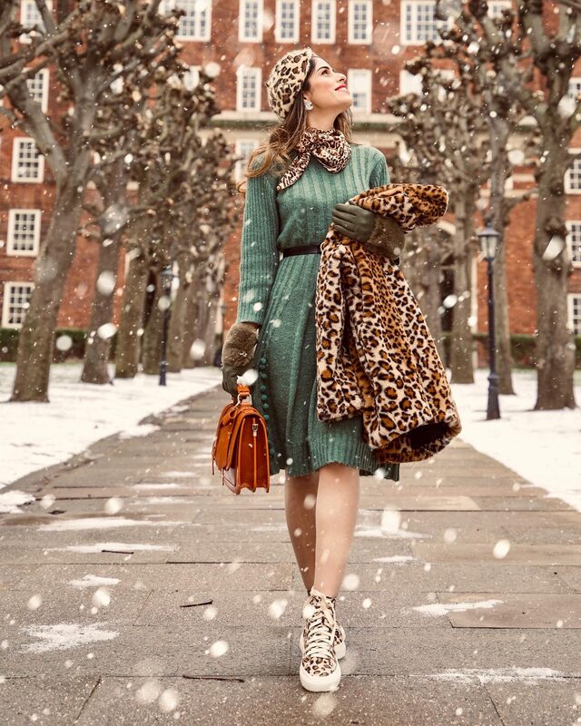 Roopsi Bansal in a green ribbed knit dress and leopard-print beret and scarf, carrying a leopard faux-fur coat over her arm, walking through falling snow on a tree-lined path