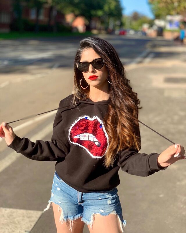 Roopsi Bansal in a black hoodie with a red sequined lip graphic and denim cutoff shorts, standing on a sunny London street