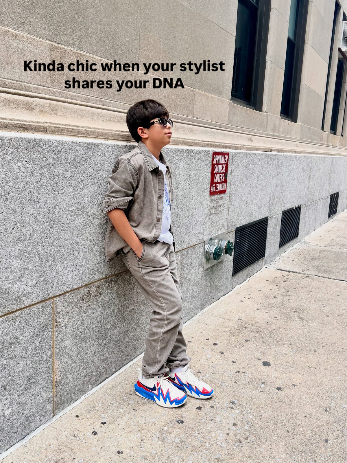 Roopsi Bansal's son leaning against a stone building in a tan denim jacket and matching pants, sunglasses on, styled by his mom, on a Manhattan sidewalk