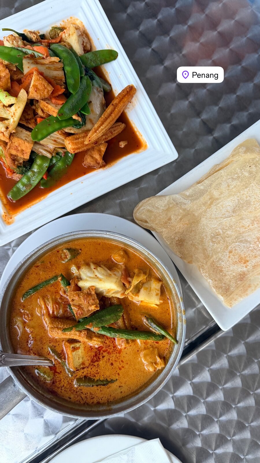 A plate of stir-fried tofu, snow peas, baby corn and cabbage in chili sauce next to a bowl of curry with roti canai flatbread, tagged Penang
