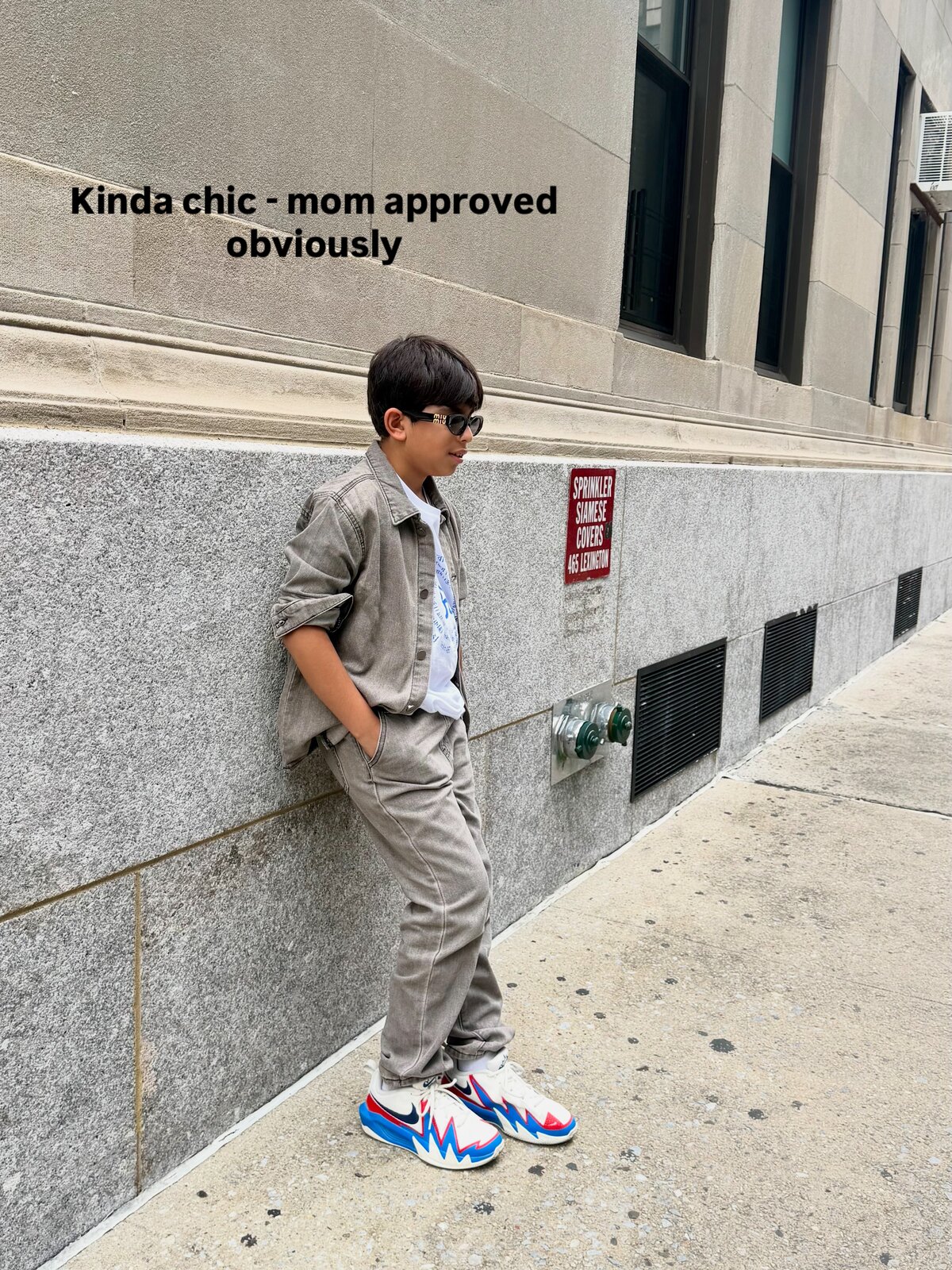 Roopsi Bansal's son in a tan denim jacket, matching pants, and sunglasses, leaning against a granite wall on a New York City sidewalk