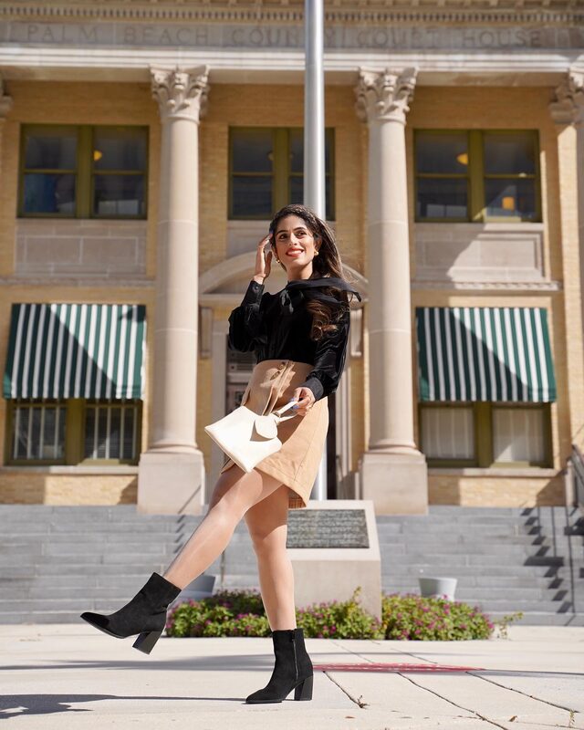 Roopsi Bansal in a black velvet blouse, camel skirt, and black ankle boots, carrying a white handbag, mid-stride in front of the Palm Beach County Courthouse