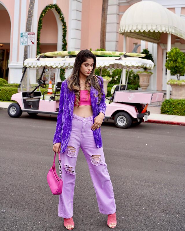 Roopsi Bansal in a crushed-velvet purple blazer over a hot pink crop top and distressed lilac jeans, standing in front of a pink golf cart with a surfboard on the roof