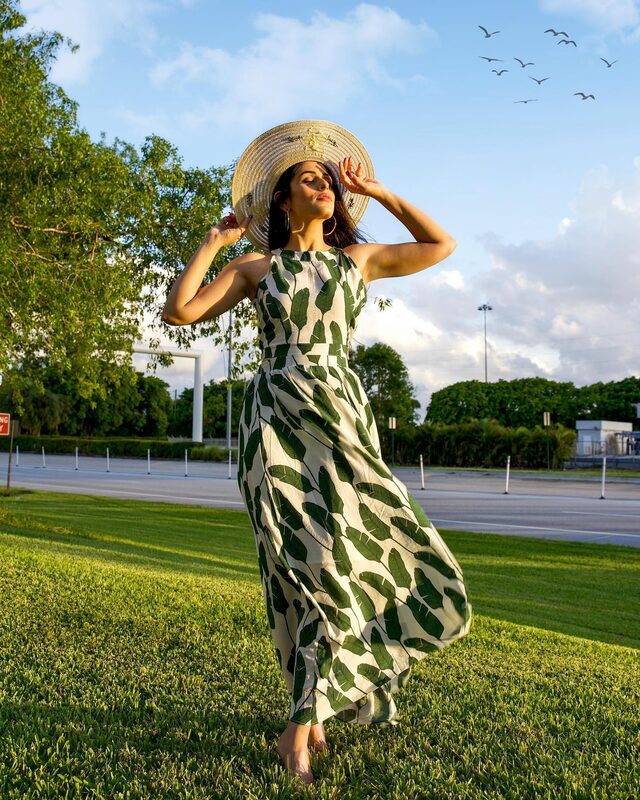 Roopsi Bansal in a green and white palm-leaf print maxi dress and a straw boater hat, standing on a lawn in the golden evening light