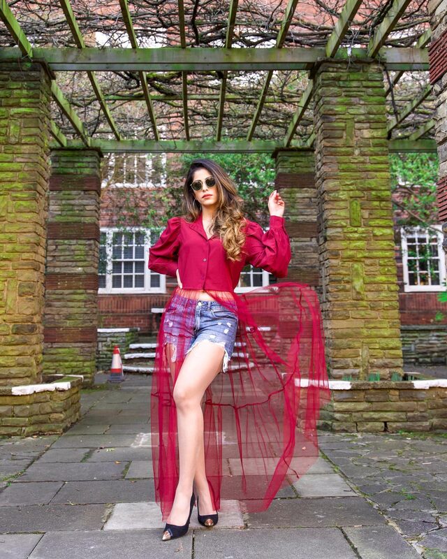 Roopsi Bansal in a maroon puff-sleeve top with a sheer tulle overlay and ripped denim shorts, posing in a mossy stone garden pergola