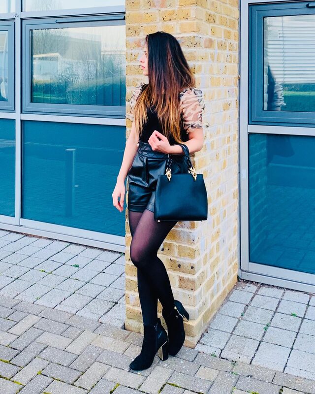 Leaning against a brick pillar in the same black organza-sleeve top and faux-leather shorts, carrying the same black chain-strap handbag