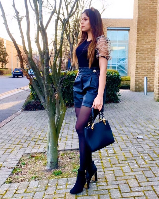 Standing on a brick pathway in a black animal-print organza-sleeve top, black faux-leather high-waist shorts, sheer tights, and ankle boots, carrying a black structured handbag with a gold chain strap