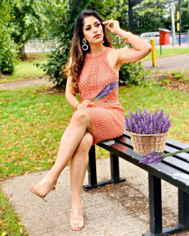 Roopsi Bansal in a coral crochet dress and statement earrings, sitting on a park bench holding a bunch of lavender