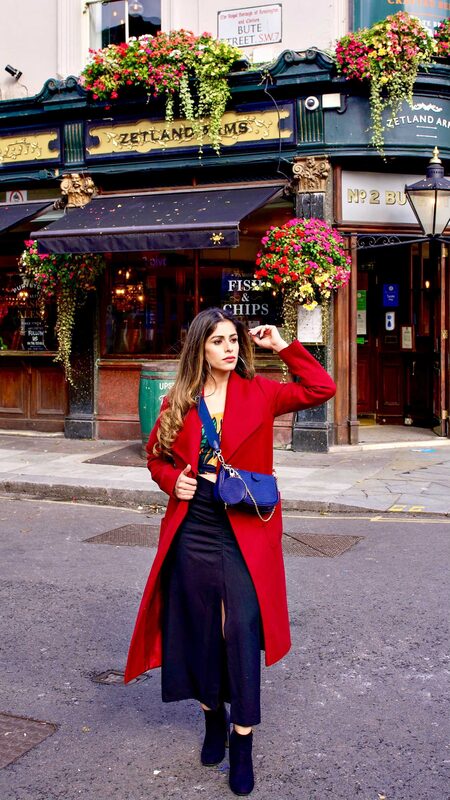 Roopsi Bansal in a red coat over a black skirt, carrying a small blue crossbody bag, standing outside a London pub with hanging flower baskets