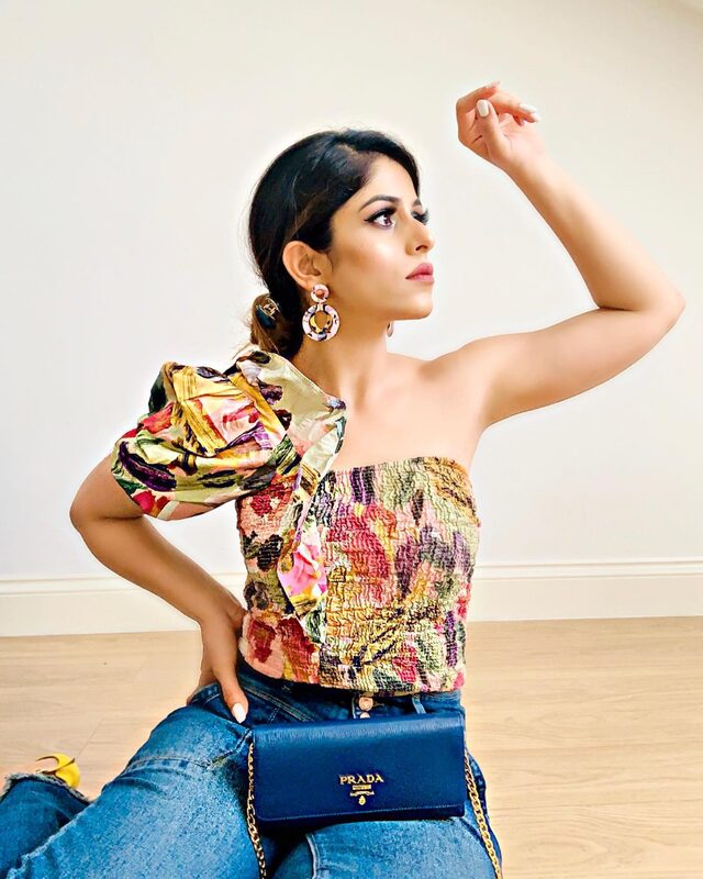 Sitting on the floor at home wearing a one-shoulder floral textured top with a puff sleeve, statement earrings, and a blue Prada bag over denim