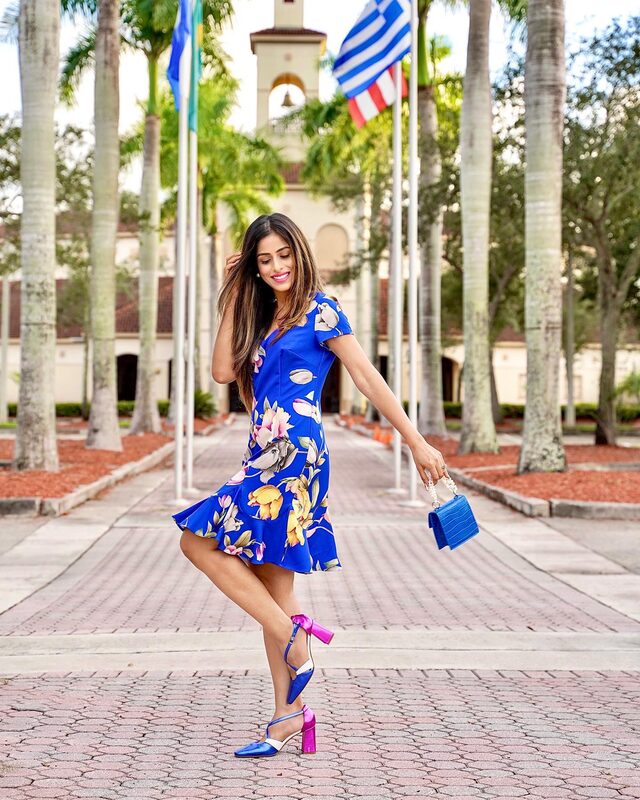 Roopsi Bansal in a cobalt blue floral-print dress with a ruffled hem, holding a blue handbag, balancing on one leg on a palm-lined promenade with flags in the background
