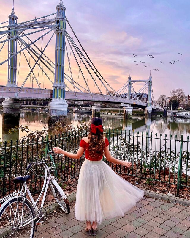 Roopsi Bansal from behind in a red velvet top and cream tulle skirt with a red bow in her hair, standing by a bicycle next to the Thames with Albert Bridge lit up at sunset