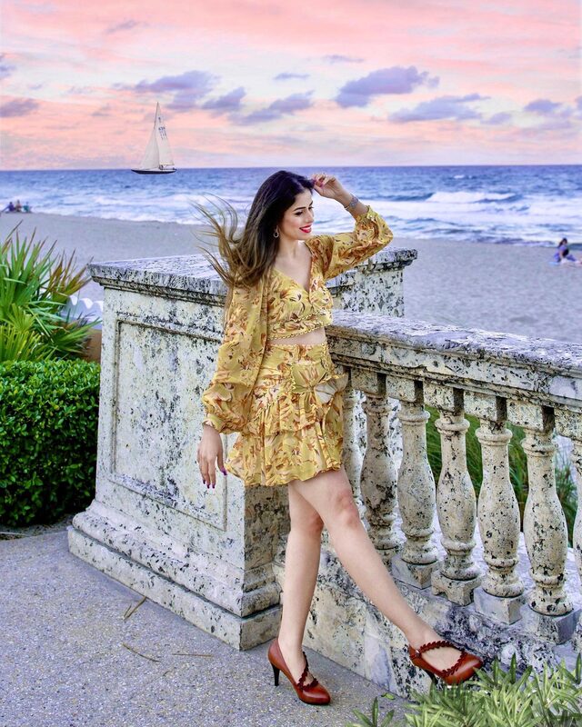 Roopsi Bansal in a yellow floral co-ord set, leaning against a stone balustrade at Palm Beach at sunset with a sailboat passing in the distance