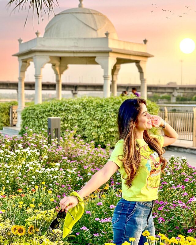 Roopsi Bansal in a neon yellow graphic tee and jeans, walking through a wildflower bed at sunset on the River Front in Lucknow, with a domed pavilion behind her