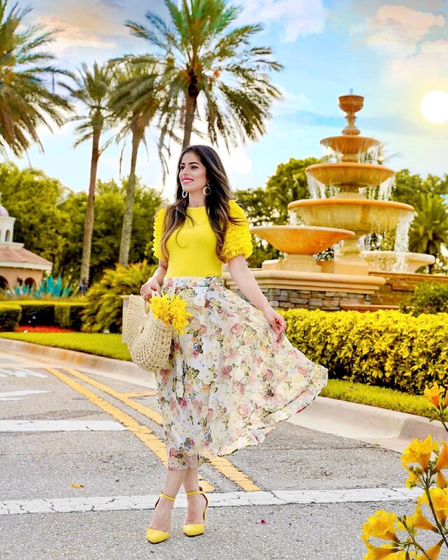 Roopsi Bansal in a yellow ruffle-sleeve top and floral flare skirt, holding a straw basket bag, in front of a tiered stone fountain in Wellington, Florida