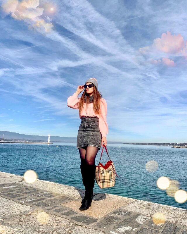 Roopsi Bansal in a pink knit sweater, grey pom-pom beanie, denim skirt and tall black boots, holding a Burberry check tote on a lakeside promenade in Switzerland