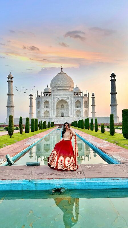Roopsi Bansal in a red and gold lehenga sitting by the reflecting pool in front of the Taj Mahal at sunset