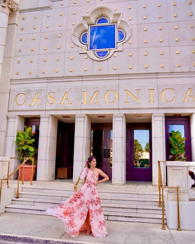 Roopsi Bansal in a blush pink one-shoulder floral maxi dress from SHEIN, standing in front of the Casa Monica hotel entrance with its gold lettering